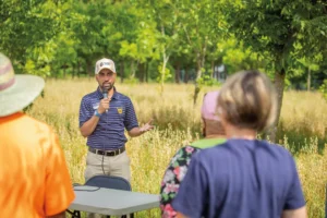 Biswanath Dari, Ph.D., a natural resource specialist with Cooperative Extension at North Carolina A&T State University, speaks into a microphone while standing in a field of tall grass. He is addressing a small group of people, some of whom are partially visible from the back. Dr. Dari is wearing a striped polo shirt with the university logo and a branded cap. The background shows a natural setting with green trees and vegetation.