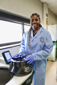 Breyana Robinson, wearing a light blue lab coat with the North Carolina A&T State University logo and purple gloves, stands beside a lab centrifuge, smiling as she holds the equipment. She is in a well-lit laboratory setting.
