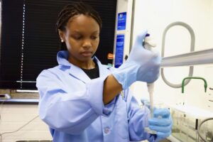 Sierra Franco, an Undergraduate Research Scholar, wearing a light blue lab coat and gloves, carefully uses a pipette in a laboratory setting. She is focused on her project related to renewable resources.