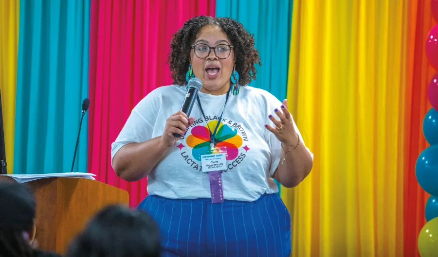 A woman with curly hair, glasses, and large turquoise earrings speaks into a microphone at a podium. She wears a white T-shirt with a colorful logo that reads