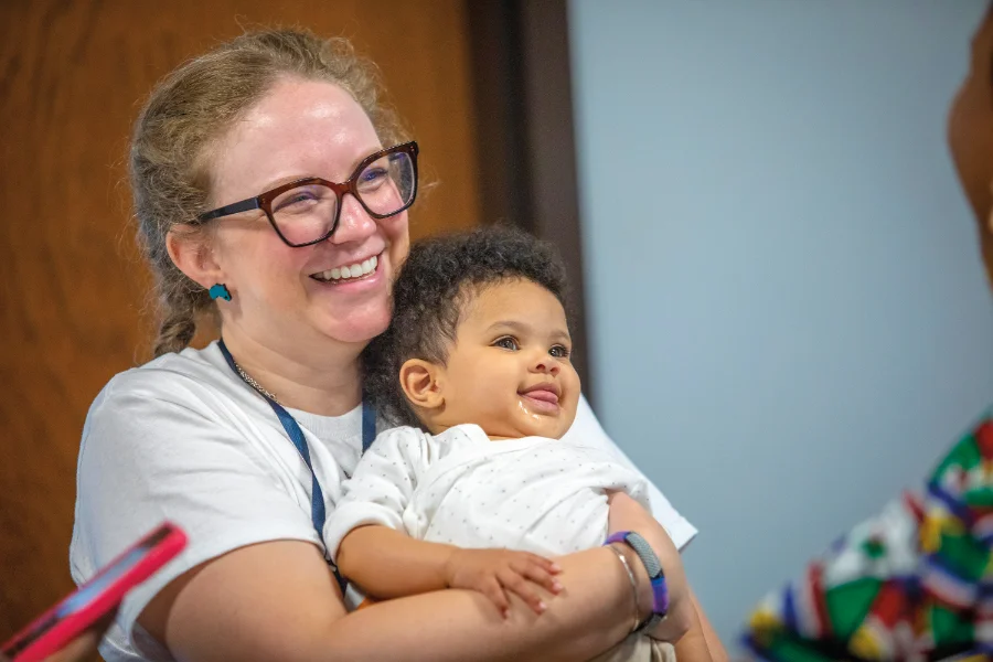 A smiling woman with glasses and blue earrings holds a baby in her arms. The baby, dressed in a white polka-dotted onesie, looks to the side with its tongue slightly sticking out. The background features a wooden door and a blurred figure wearing a colorful patterned outfit.