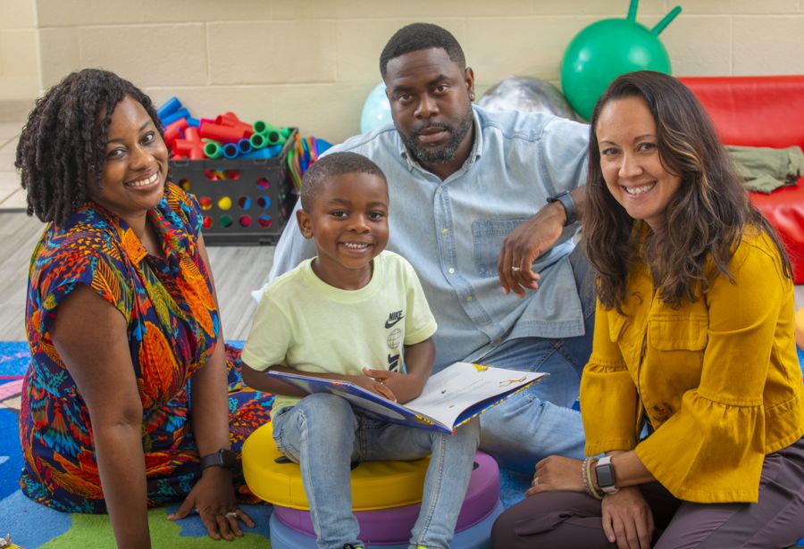 A young boy with a bright smile sits on a colorful stool, holding an open book, surrounded by three adults in a playroom. The woman on the left wears a vibrant patterned dress and smiles warmly. The man in the center wears a light blue denim shirt and has a serious yet attentive expression. The woman on the right wears a mustard-yellow jacket and smiles. The background features play equipment, including foam rollers, a red couch, and a green exercise ball.